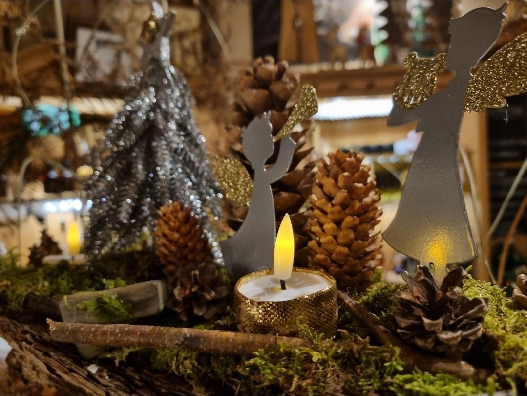 Eine Weihnachtsszene mit einer brennenden Kerze, Tannenzapfen, einer Moosbasis und Engelfiguren. Ein silberner Baum steht dahinter.