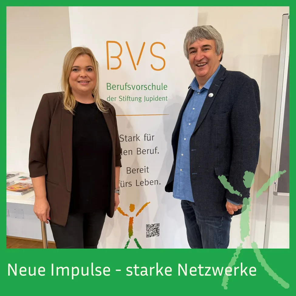 Zwei Personen, eine Frau und ein Mann, stehen vor einem Banner mit dem Text 'BVS Berufsvorschule der Stiftung Judipent'. Die Frau lächelt und hält einen Stock, während der Mann lächelt und in die Kamera schaut. Das Banner hat einen QR-Code darauf.
