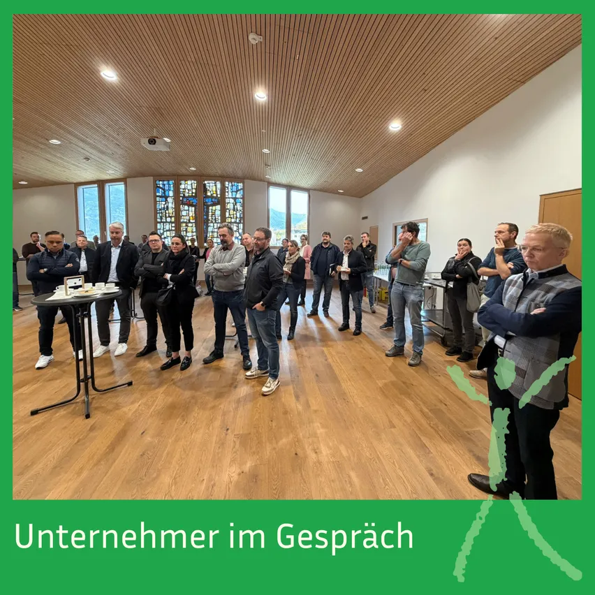 Eine Gruppe von Menschen steht in einem Raum, möglicherweise in einem Gespräch. Der Raum hat Holzböden und eine Decke mit Lichtern. Einige Menschen haben die Arme verschränkt, während andere etwas betrachten. Der Raum hat auch Fenster mit Buntglas.