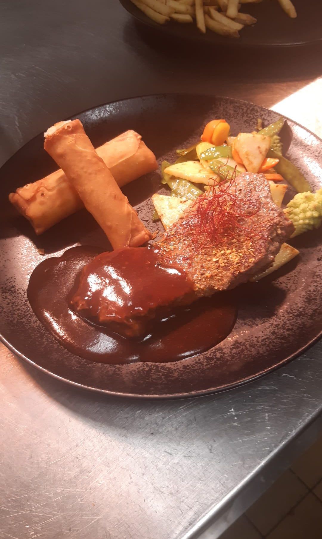 Ein Teller mit Steak in Soße, garniert mit roten Fäden, begleitet von einer Auswahl an Gemüse wie Brokkoli, Karotten und grünen Bohnen sowie zwei Frühlingsrollen.