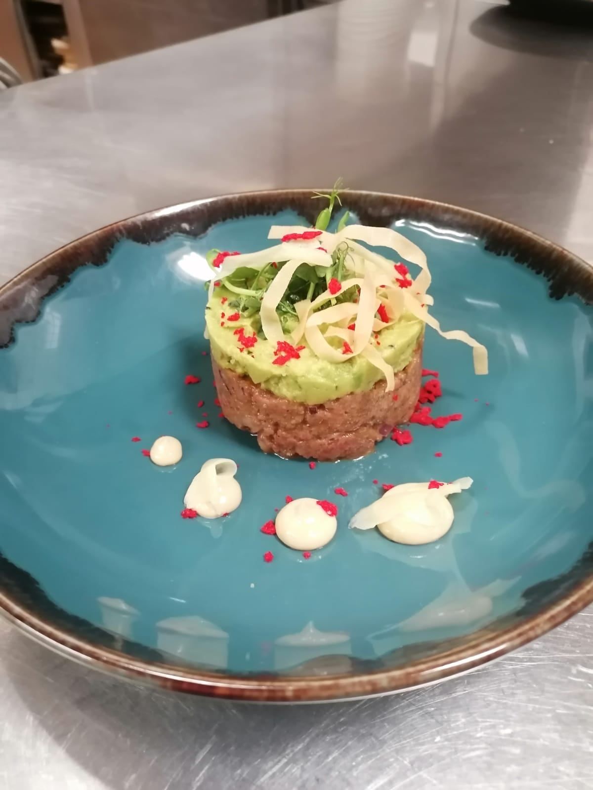 A gourmet dish on a blue plate, topped with avocado, white cream, and red and white garnishes.