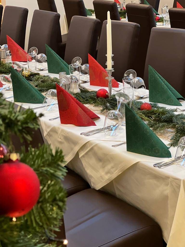 Eine festliche Tischdekoration mit roten und grünen Servietten, Weihnachtskugeln und einer Kerze auf einem silbernen Halter. Der Tisch ist mit einer weißen Tischdecke bedeckt.