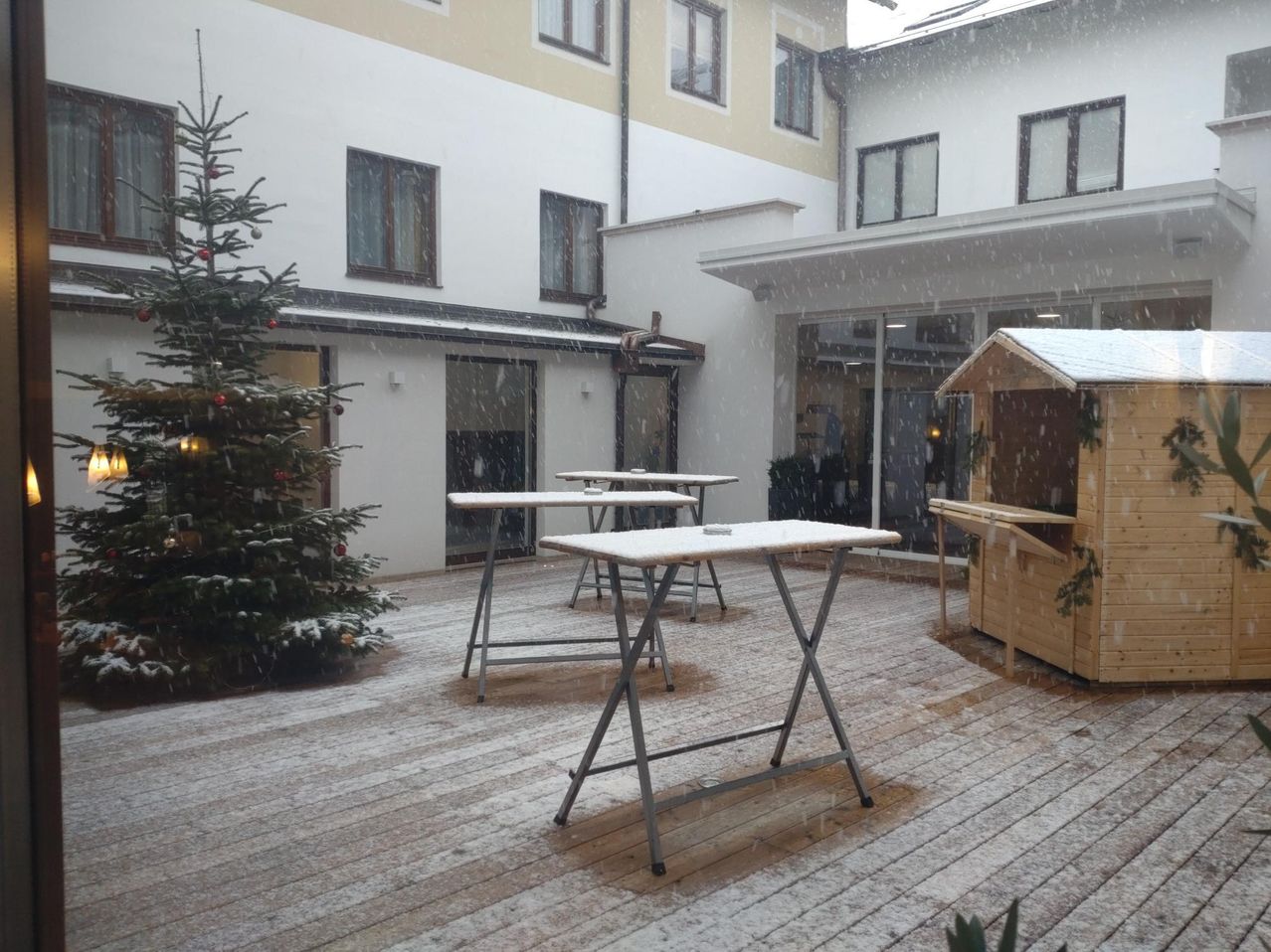 Ein Patiobereich mit einem Weihnachtsbaum, einigen Tischen und einem kleinen Holzhäuschen ist zu sehen. Der Patio ist von einem Gebäude mit mehreren Fenstern umgeben. Es schneit.