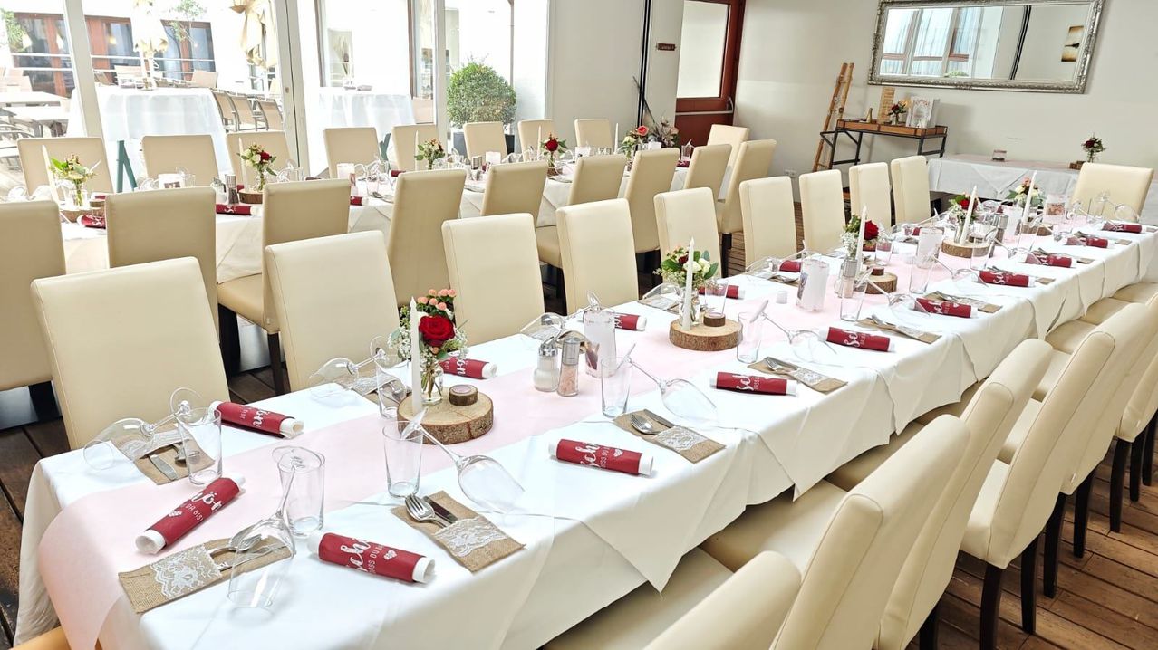 Ein großer Speisesaal mit vielen Stühlen und Tischen, alle für eine besondere Veranstaltung vorbereitet. Jeder Tisch hat Blumen, Kerzen und rote Servietten. Der Raum ist gut beleuchtet und geräumig.