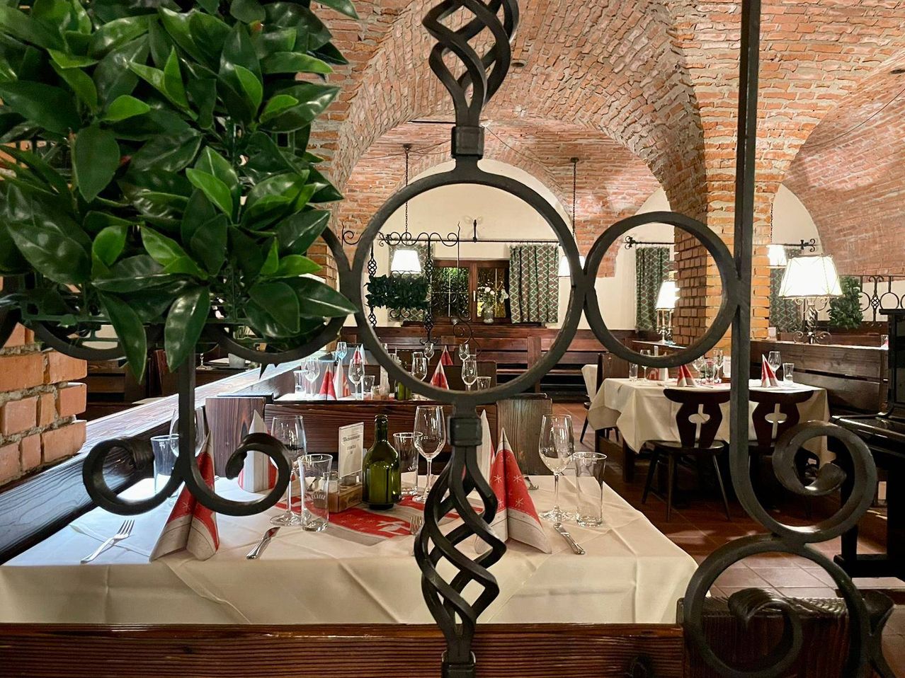 A restaurant with a brick ceiling and a table set for a meal. A bottle, wine glasses, and napkins are on the table. A plant and a lamp hang from the ceiling.