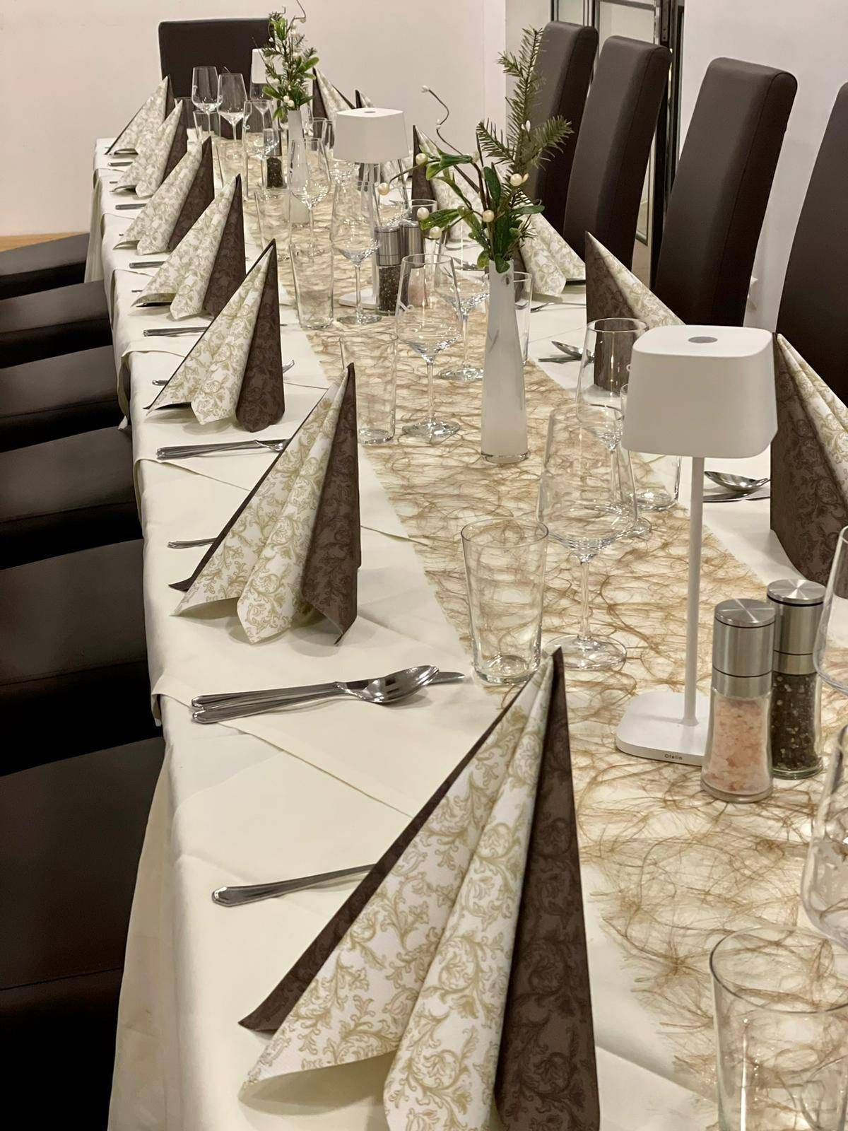 A long dining table set for a formal event with white tablecloth, folded napkins, wine glasses, and utensils. There is a white vase with flowers, and salt and pepper shakers on the table.