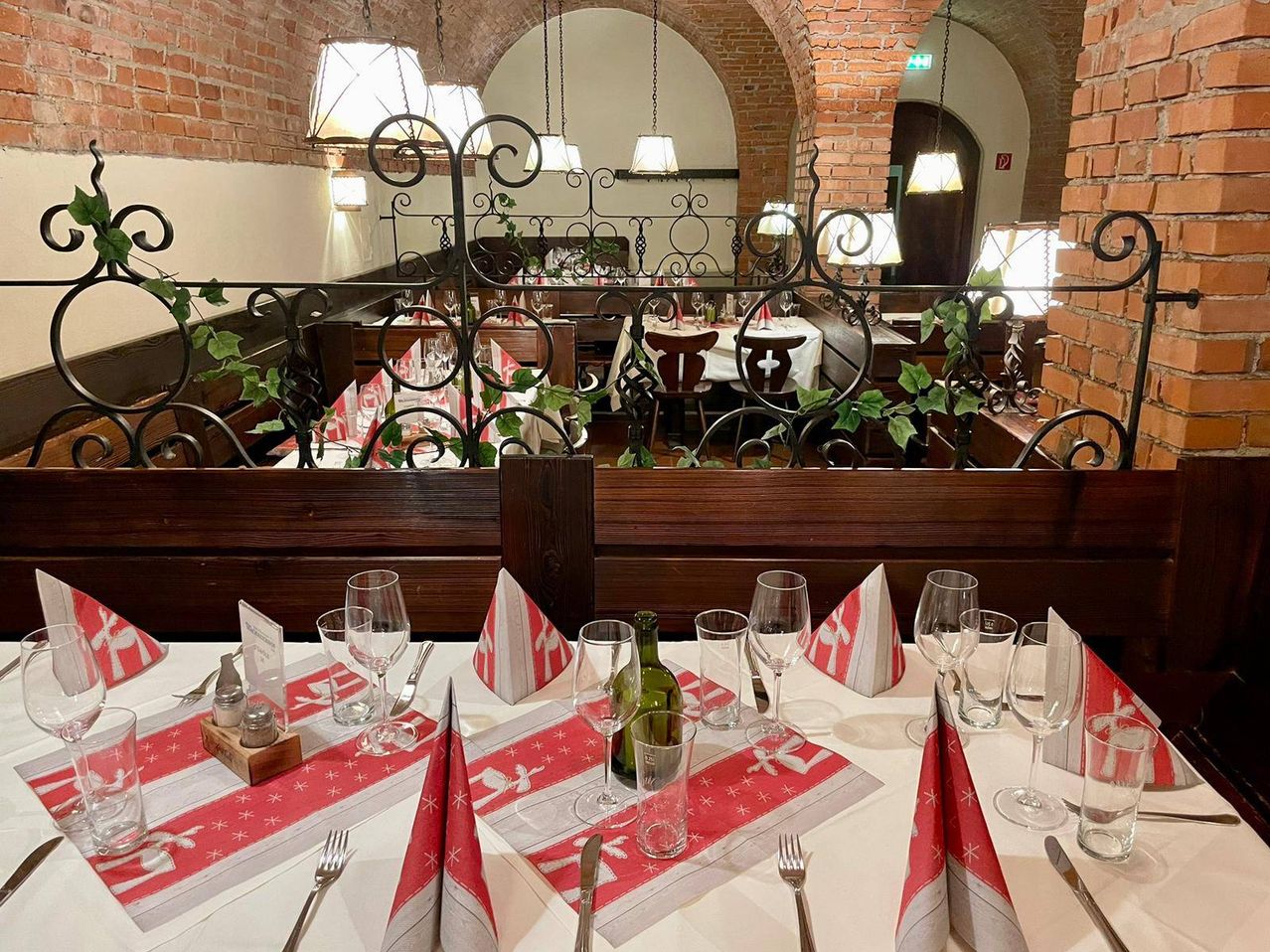Ein Restauranttisch, der für eine Mahlzeit mit Gläsern, Besteck und einer Weinflasche gedeckt ist. Der Tisch ist mit einer rot-weißen Tischdecke bedeckt. Dahinter sind eine Ziegelwand und hängende Lampen zu sehen.