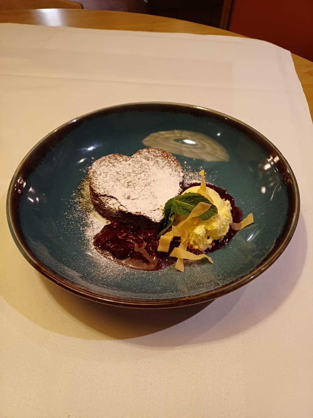 Ein herzförmiges Schokoladen-Dessert auf einem türkisen Teller, garniert mit Puderzucker, Eiscreme und Kirschsauce.