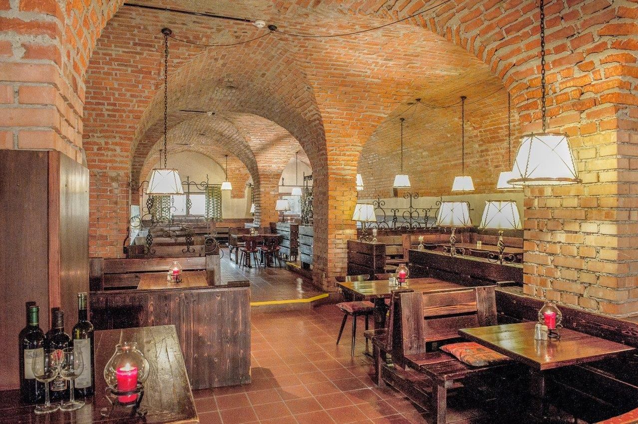 Ein Restaurant mit Ziegelwänden, gewölbten Decken und Holzmöbeln. Tische und Stühle sind aufgestellt, und Lampen hängen von der Decke. Der Boden ist gefliest, und es gibt eine gelbe Linie auf dem Boden.