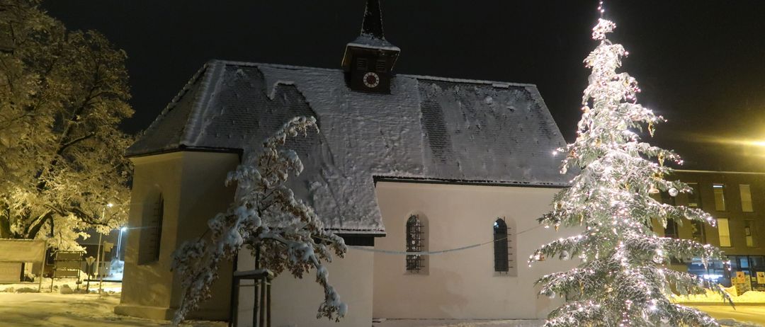 Eine Kirche mit einem Turm und einer Uhr steht im Schnee, beleuchtet von ihrem eigenen Licht. Ein geschmückter Weihnachtsbaum steht in der Nähe, ebenfalls beleuchtet.