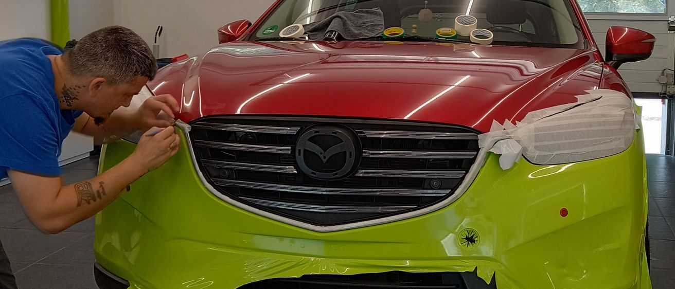 Ein Mann arbeitet in einer Garage an einem roten und grünen Auto. Das Auto ist in Plastik eingewickelt und hat ein Mazda-Logo. Die Garage hat weiße Wände, Lampen und Fenster.