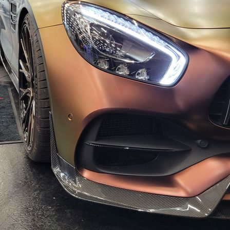 Ein Nahaufnahme des goldenen Sportwagens mit Frontstoßstange und Scheinwerfern, im Hintergrund ein schwarzer Teppich.