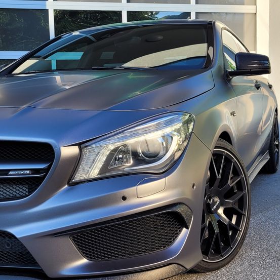 Ein schlanker, silberner Mercedes-AMG CLA 45 ist vor einem Glasgebäude geparkt und zeigt seinen aggressiven Frontgrill und schwarze Leichtmetallräder.