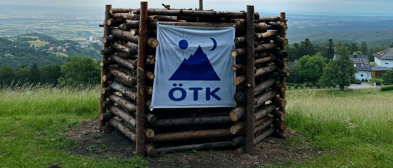 Ein Holzkreuz mit einem Banner hängt oben auf einem Stapel Holzscheite, wahrscheinlich auf dem Gipfel eines Hügels, mit Blick auf das Tal unten.