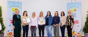 Sieben Frauen stehen in einer Reihe und lächeln für ein Foto. Sie befinden sich in einem Raum mit einem roten Teppich und zwei Bannern hinter ihnen. Sie halten Urkunden in den Händen, und zwei von ihnen halten größere gerahmte Urkunden.