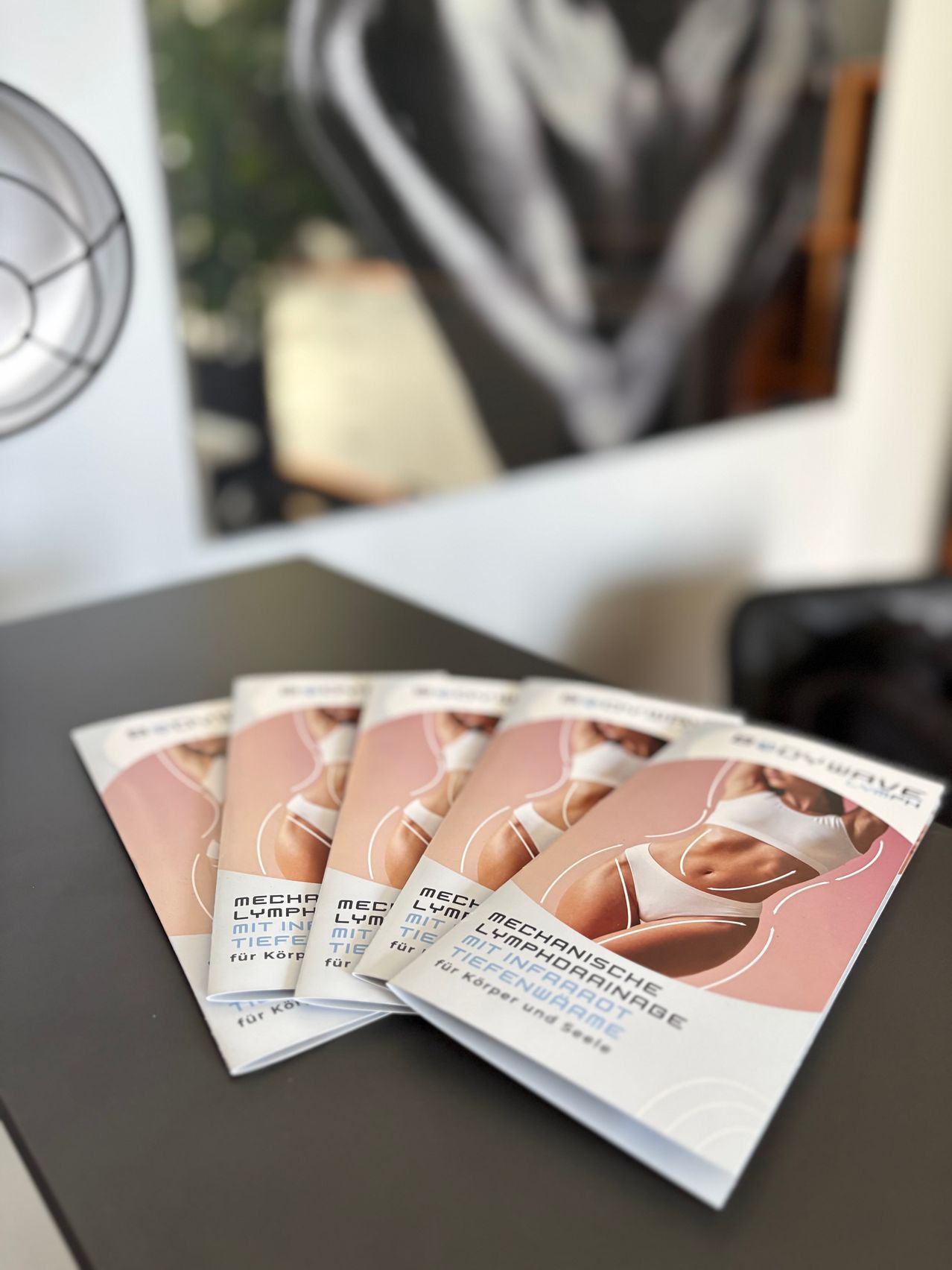 Drei Broschüren über mechanische Lymphdrainage liegen auf einem Tisch. Die Broschüren haben rosa und weiße Umschläge mit Bildern von Frauen in weißer Unterwäsche.