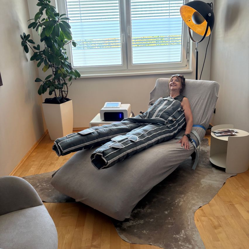 Eine Frau liegt auf einem Liegestuhl mit einer Decke, lächelt und schaut in die Kamera. Es gibt eine Pflanze und eine Lampe im Raum.