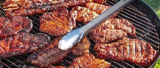 Eine Person verwendet eine Grillzange, um Fleisch auf einem Grill im Freien zu wenden. Der Grill ist voll mit Steaks und Spießen.