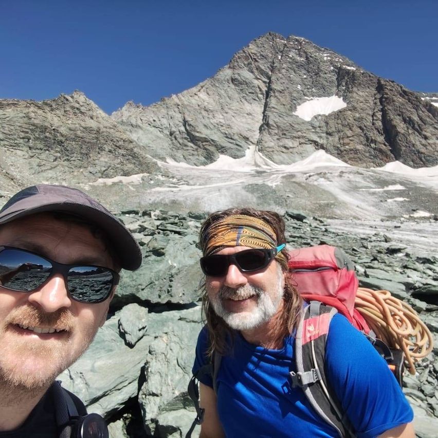 Zwei Männer in Kletterausrüstung lächeln vor einer felsigen verschneiten Berg.