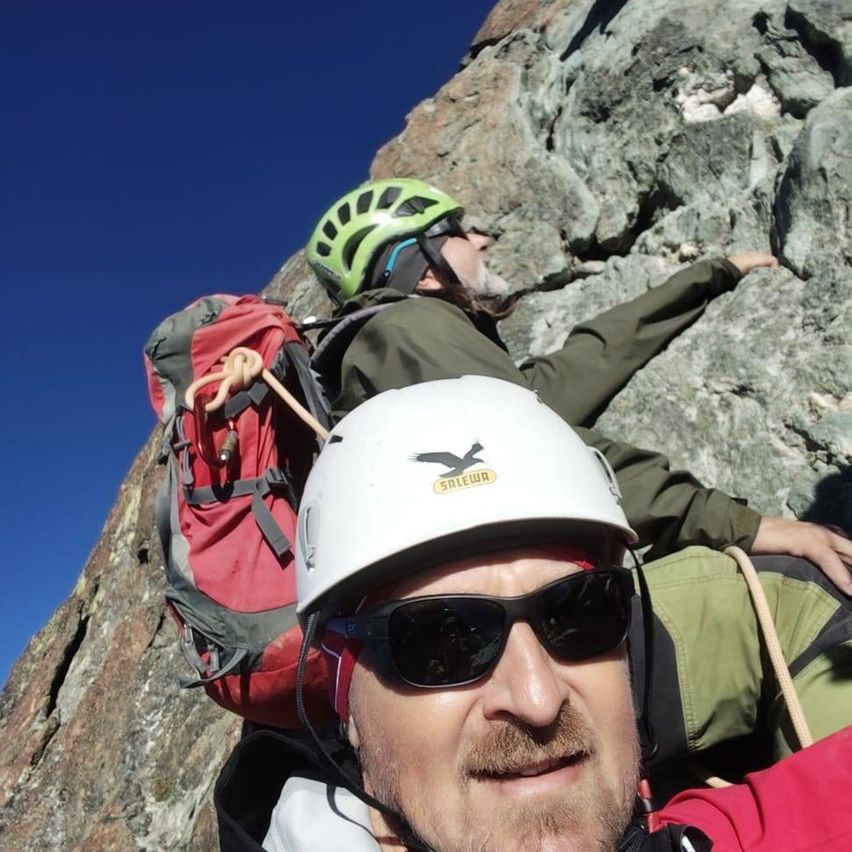 Zwei Männer klettern einen felsigen Berg, beide tragen Helme und Rucksäcke. Der Mann vorn trägt eine Sonnenbrille und einen weißen Helm. Der Mann dahinter trägt einen grünen Helm.