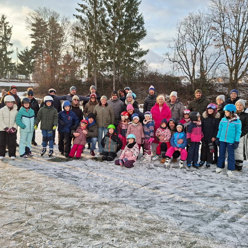 Eine Gruppe von Menschen steht und sitzt auf einem verschneiten Feld. Sie tragen Winterkleidung und einige tragen Helme. Bäume und ein Haus sind im Hintergrund.