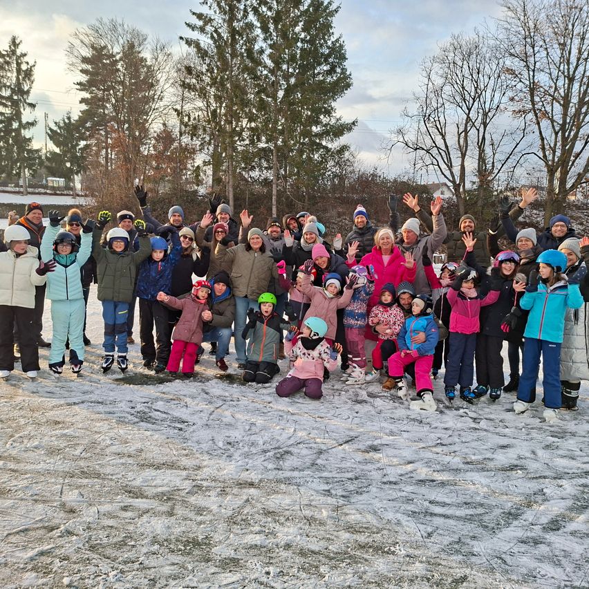 Eine Gruppe von Menschen, sowohl Erwachsene als auch Kinder, lächelt und posiert für ein Foto auf einer Eisbahn. Sie tragen Winterkleidung und einige haben Skiausrüstung. Im Hintergrund sind Bäume zu sehen.