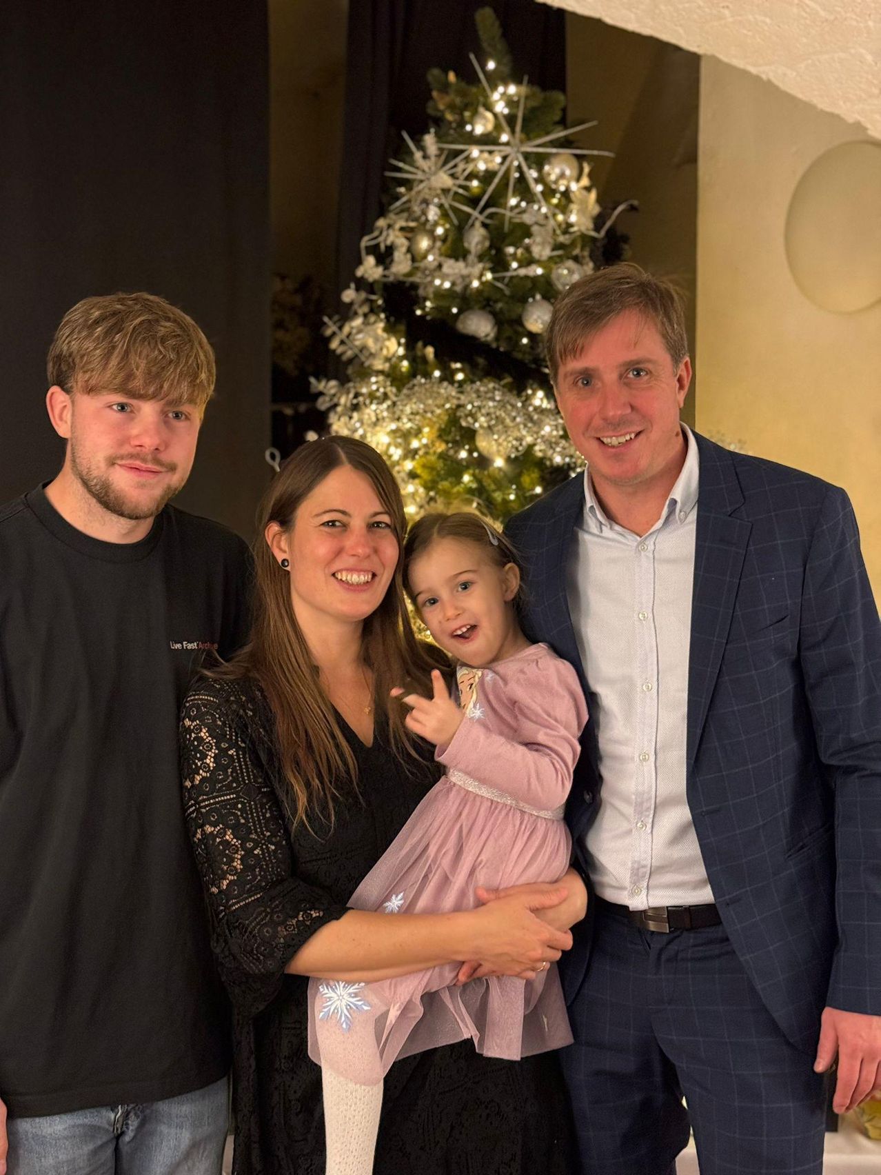 Eine vierköpfige Familie posiert für ein Foto vor einem geschmückten Weihnachtsbaum. Vater und Mutter halten ein junges Mädchen, das zur Kamera lächelt. Der Junge steht neben ihnen, alle mit festlichen Lächeln.