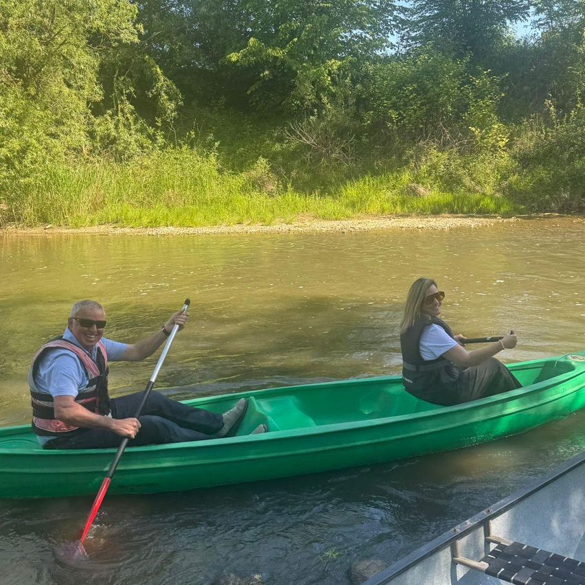 Zwei Personen genießen eine Flussfahrt in einem grünen Kanu und paddeln entlang der Wasseroberfläche. Eine Person trägt einen Schwimmweste, während die andere ein Paddel hält.