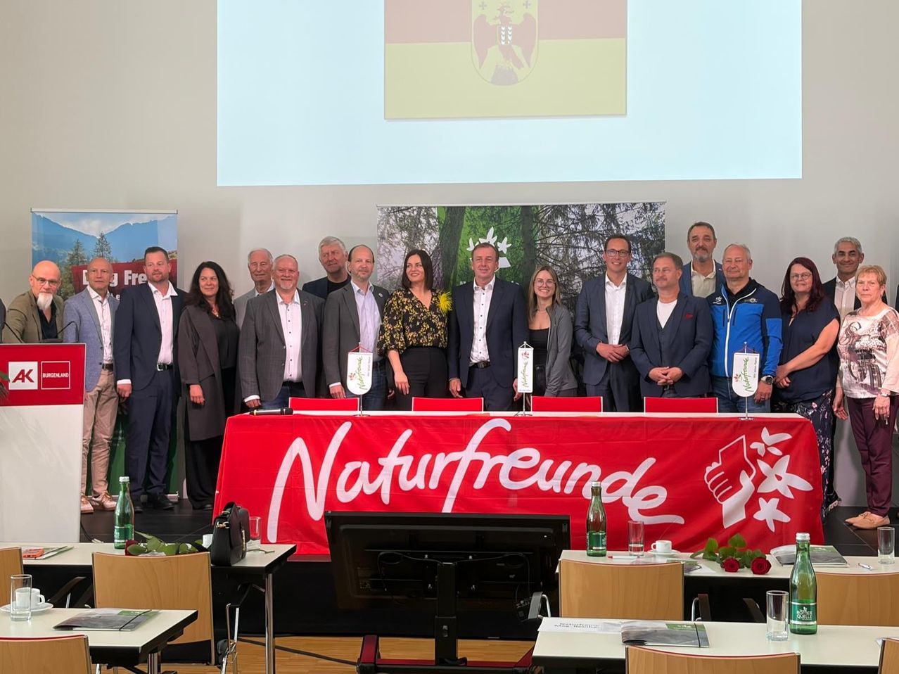 Eine Gruppe von Menschen in Anzügen und Jacken steht hinter einem Tisch mit einem roten Banner, das Naturfreunde sagt. Im Hintergrund befindet sich eine Projektionswand.