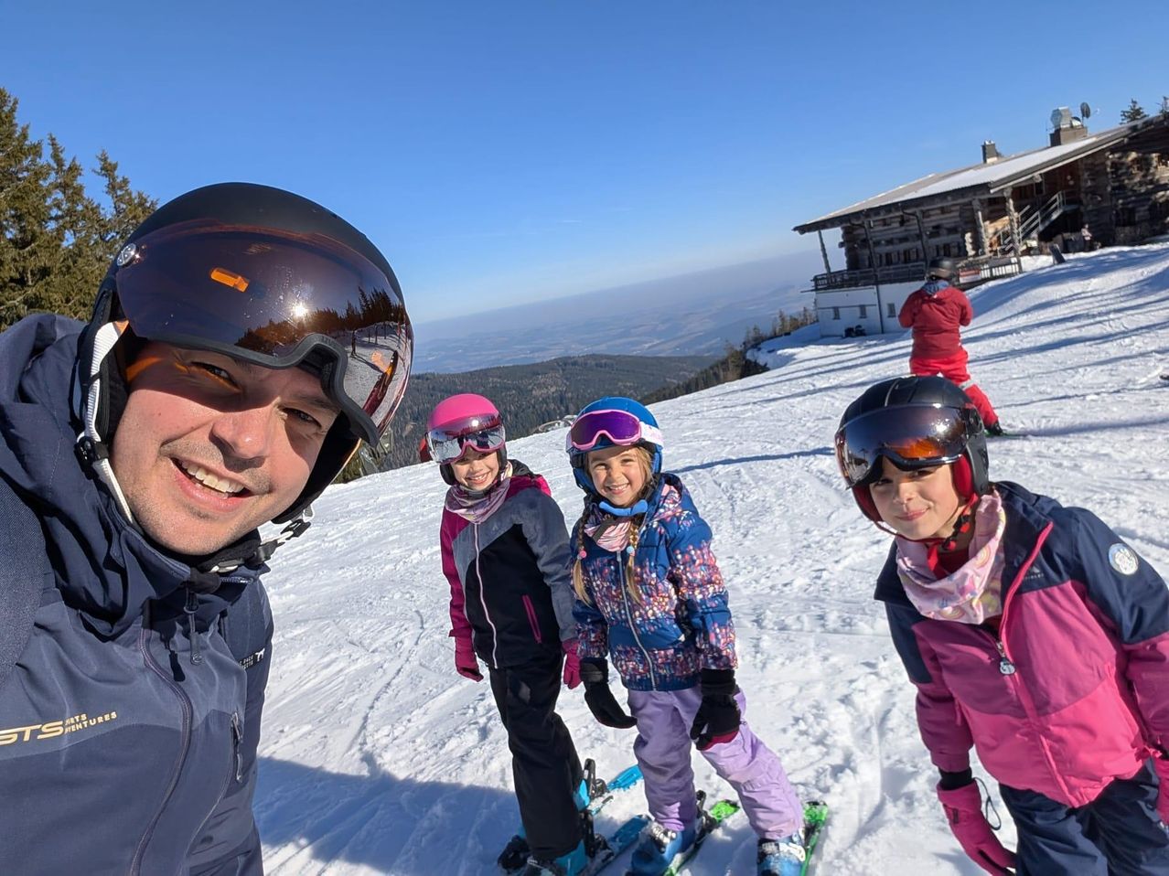Ein Mann und drei Kinder stehen auf einem verschneiten Hang und skifahren, alle tragen Winterkleidung und Helme. Im Hintergrund ist ein Chalet zu sehen, neben dem eine Person steht.