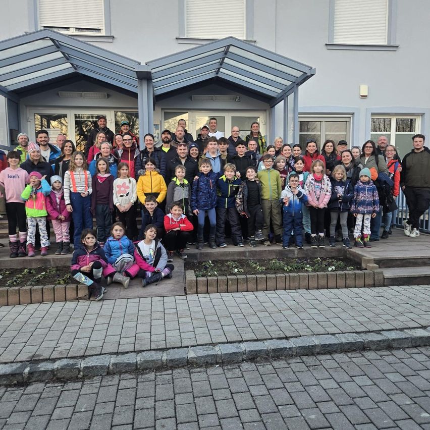 Eine Gruppe von Menschen, einschließlich Kindern, posiert für ein Foto vor einem Gebäude mit mehreren Fenstern und einem überdachten Eingang.