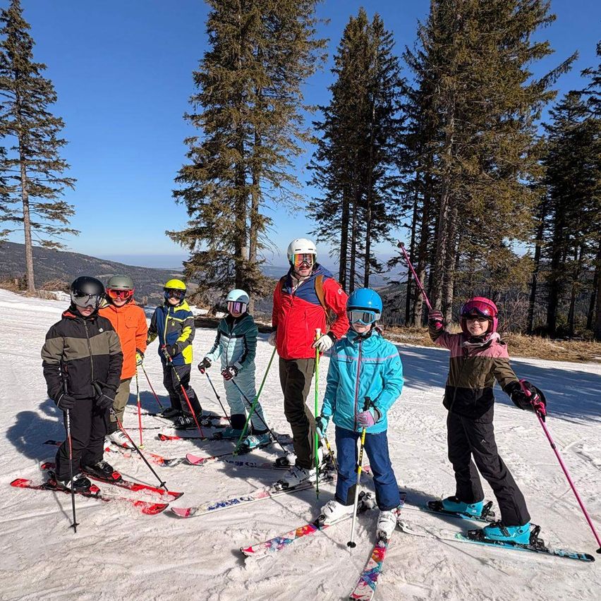 Eine Gruppe von Skifahrern und Snowboardern posiert für ein Foto auf einem verschneiten Berg mit Kiefern im Hintergrund.