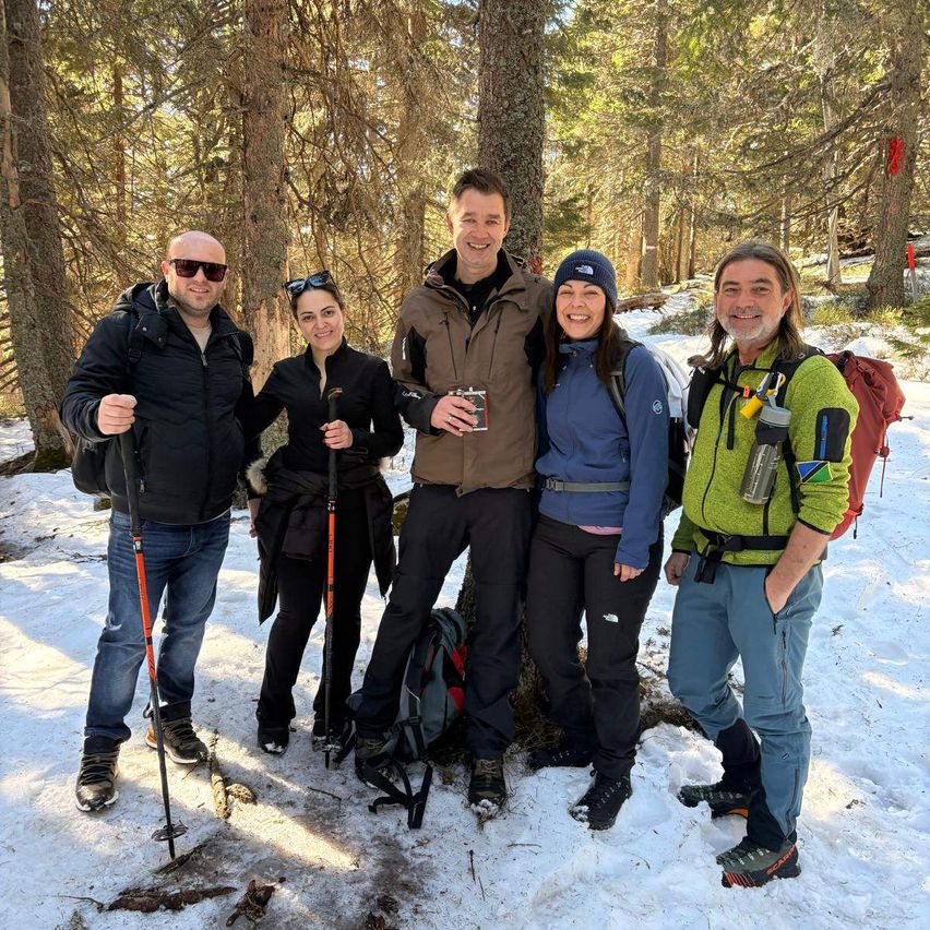 Fünf Personen in Winterkleidung stehen in einem verschneiten Bereich, umgeben von Bäumen. Sie lächeln und posieren für ein Foto.