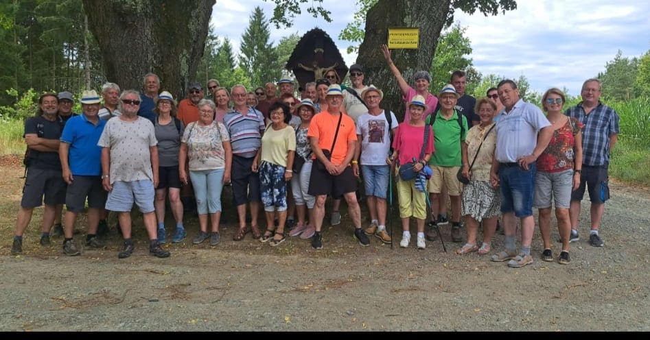 A group of people, dressed in casual summer attire, are standing in a line in front of a forest backdrop. Some are wearing hats and sunglasses. A yellow sign is visible on a tree behind them.