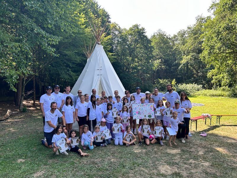 Eine Gruppe von Menschen, einschließlich Erwachsener und Kindern, versammelt sich in einem Park. Sie stehen vor einem Tipi, halten Urkunden und lächeln. Im Hintergrund befinden sich Bäume und eine Rasenfläche.