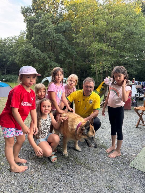 Eine Gruppe von Kindern und ein Mann stehen in einem Waldgebiet mit einem Hund. Der Mann kniet und hält den Hund, während die Kinder um ihn herum stehen.