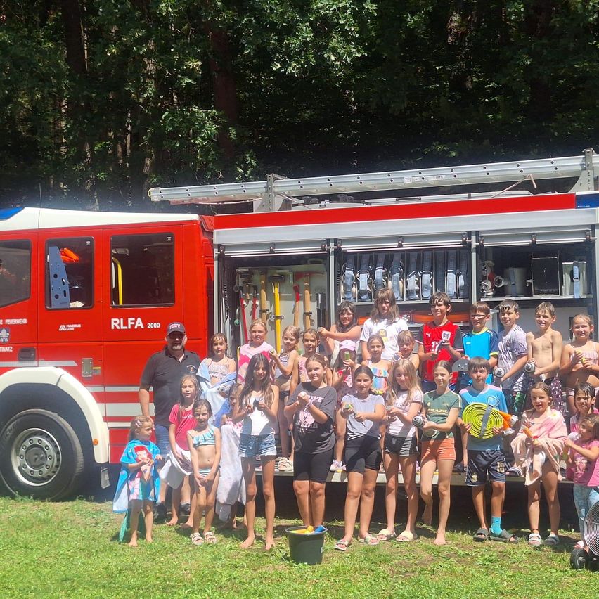 Eine Gruppe von Kindern und Erwachsenen steht vor einem roten Feuerwehrauto, einige halten Wasserpistolen und Handtücher, ein Mann in einem Hut ist in der Nähe.