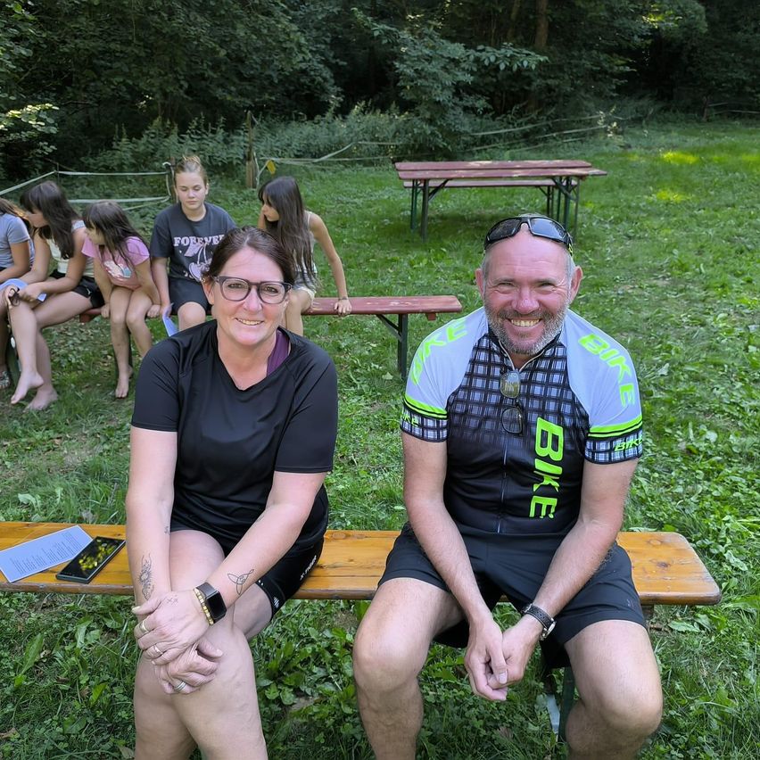 Ein Mann und eine Frau sitzen lächelnd auf einer Parkbank. Hinter ihnen sitzen eine Gruppe von Kindern auf Bänken und Tischen. Der Mann trägt ein Radtrikot und die Frau ein schwarzes Shirt.