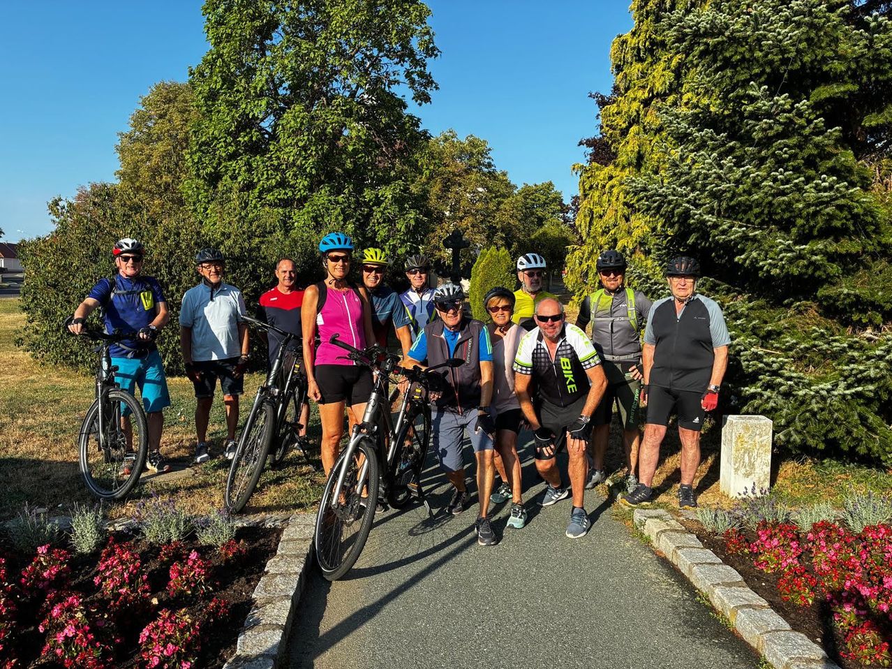 A group of cyclists in cycling gear pose for a photo in a park with lush greenery and flowers in the background.