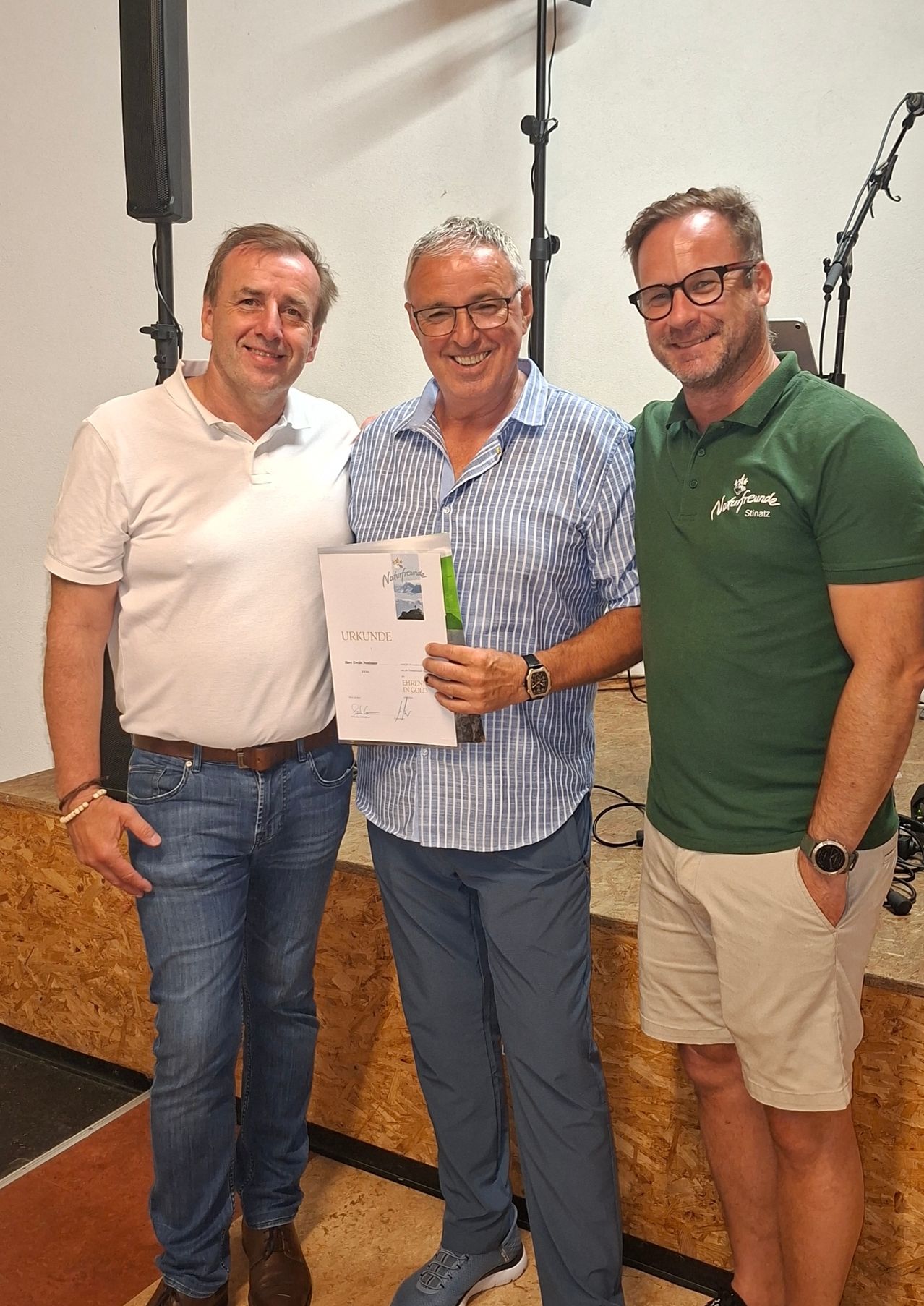 Drei Männer stehen lächelnd in einem Studio zusammen. Der Mann in der Mitte hält ein Zertifikat, die anderen flankieren ihn. Ein Holztisch und Mikrofone sind hinter ihnen.