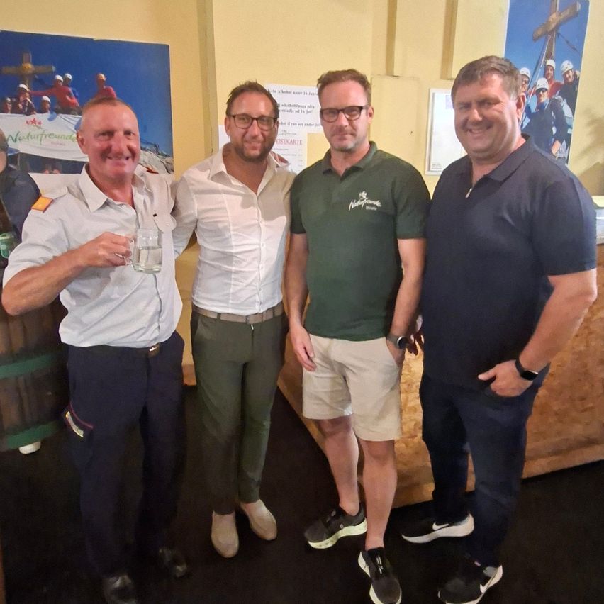 Vier Männer posieren für ein Foto. Einer hält ein Glas. Sie stehen in der Nähe einer Wand mit einem Poster und einem Fass.