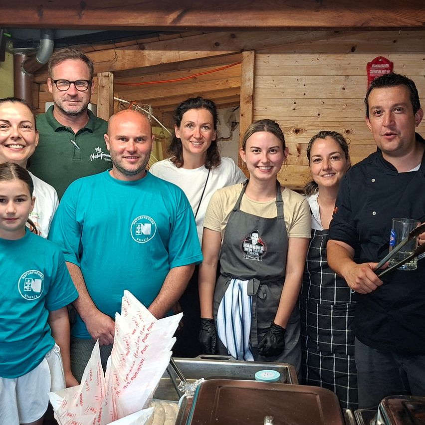 Eine Gruppe von Menschen steht vor einer Holzwand und lächelt. Ein Mann in einem schwarzen Hemd hält ein Glas und Zange. Eine Frau in einem weißen Hemd hält einen Tablett. Ein junger Junge in einem grünen Hemd hält einen Stapel Papiertüten.