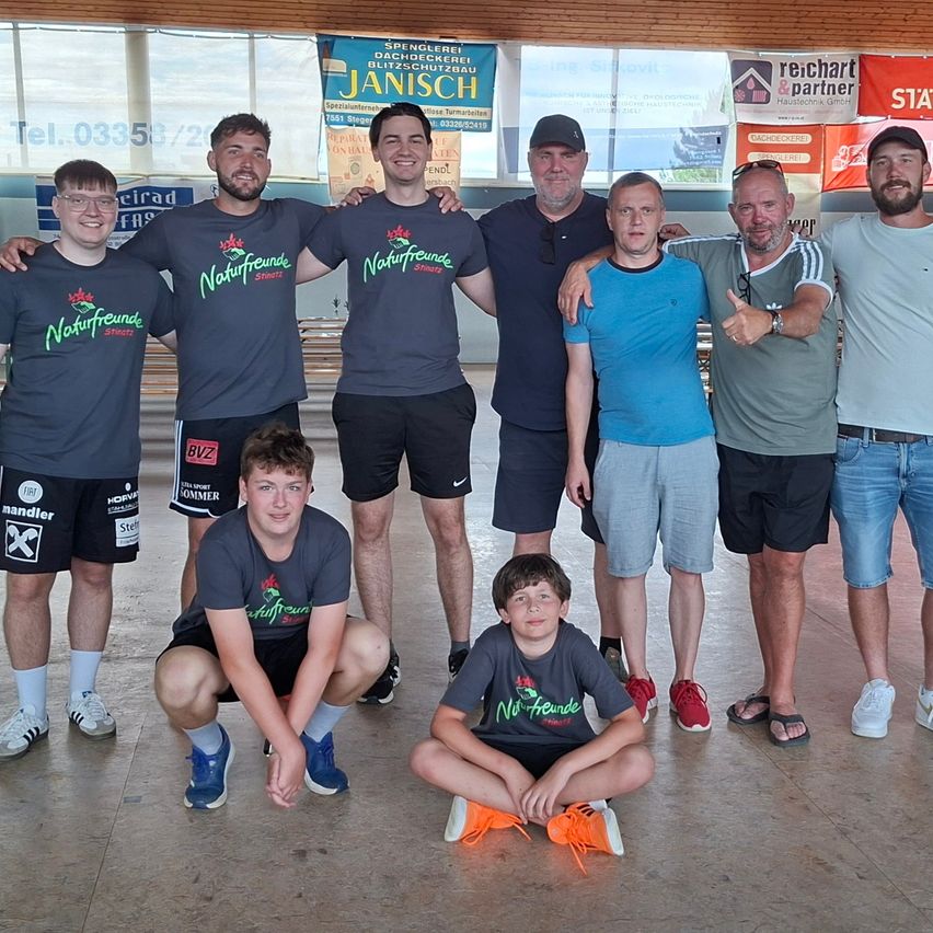 A group of people, including adults and children, are posing for a photo in a sports hall, wearing t-shirts with green prints. They are standing and sitting on the floor.