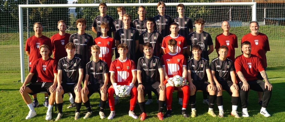 Eine Fußballmannschaft posiert für ein Gruppenfoto vor einem Tornetz, Spieler in roten und schwarzen Trikots, einige halten Fußballbälle.