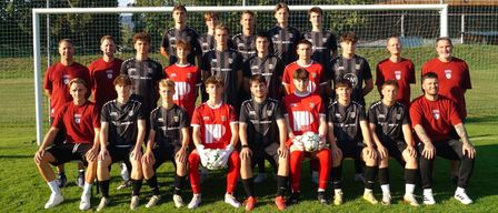 Eine Fußballmannschaft posiert für ein Gruppenfoto vor einem Tornetz, Spieler in roten und schwarzen Trikots, einige halten Fußballbälle.