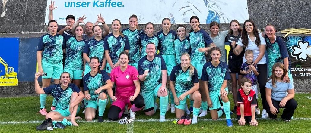 Eine Fußballmannschaft posiert für ein Foto auf einem Feld mit einem Banner und einer Anzeigetafel im Hintergrund.