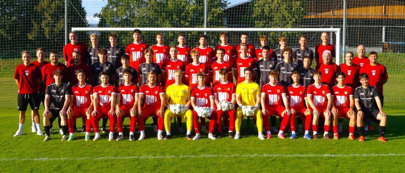 Eine Fußballmannschaft in roten und gelben Trikots posiert für ein Gruppenfoto auf einem grünen Rasen mit einem Tornetz im Hintergrund.