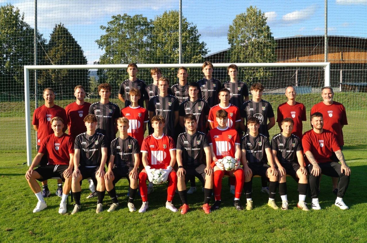 Eine Fußballmannschaft posiert für ein Foto auf einem Feld mit einem Tornetz im Hintergrund. Die Spieler tragen rote und schwarze Trikots. Einige halten Fußballbälle.
