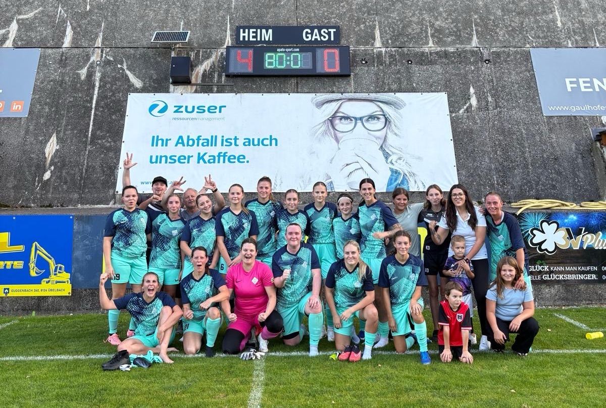 Eine Frauenfußballmannschaft posiert für ein Foto vor einem Banner mit einer digitalen Anzeigetafel, die den Spielstand 4-0 anzeigt.