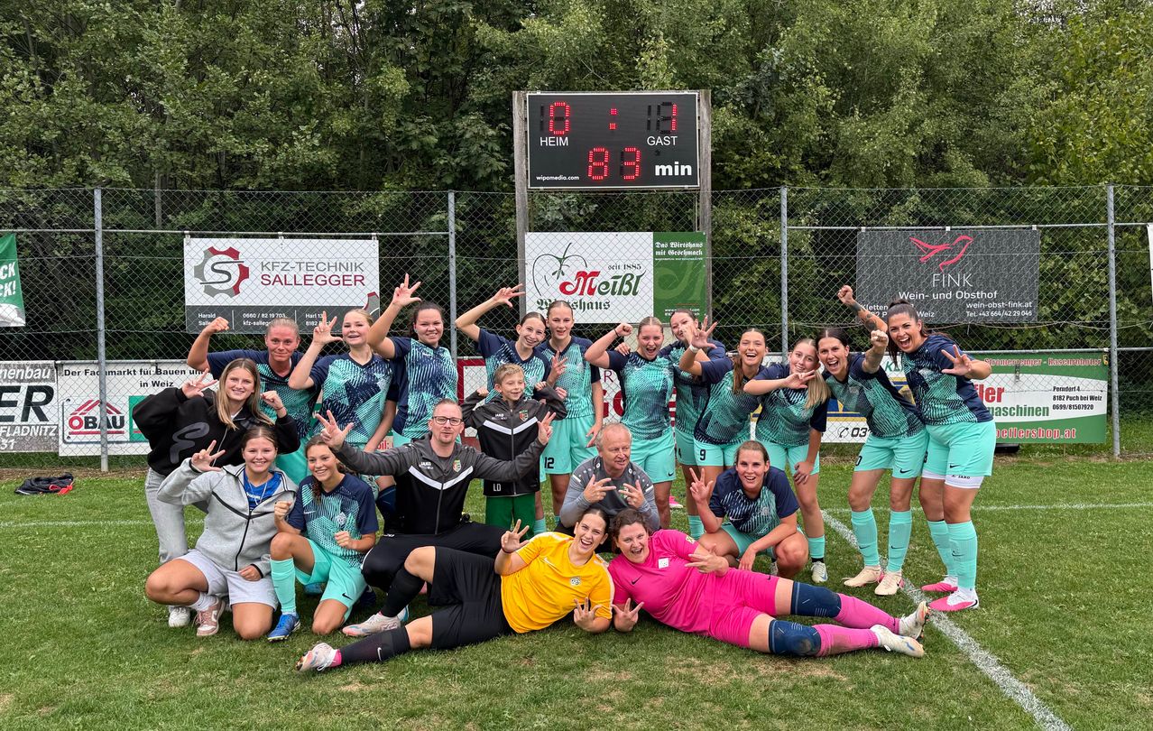 A soccer team in green and blue uniforms poses for a group photo on a field with a scoreboard in the background.